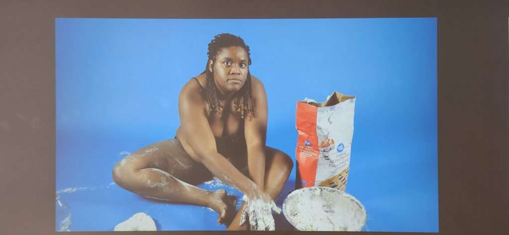 Video wall projection in which the artist sits on the floor partially dressed, making dough on the ground and her body. Detail from Racquel Rowe's exhibition titled 
