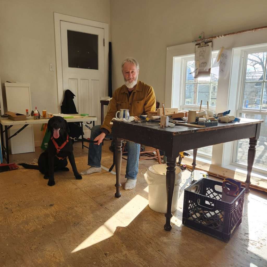 Ken Flett sits with his dog at a sunny window with his marionette carving tools and materials