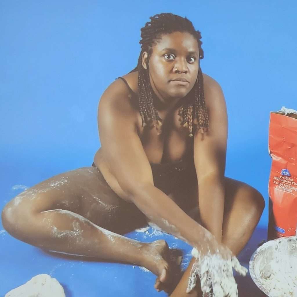 Video wall projection in which the artist sits on the floor partially dressed, making dough on the ground and her body. Detail from Racquel Rowe's exhibition titled "The Chicken is Just Dead First."