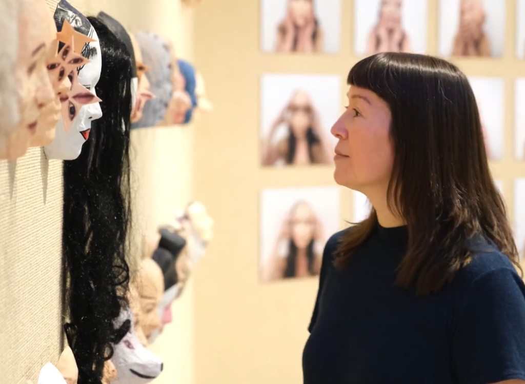 Miya Turnbull in profile looking into the eyes of masks she has created based on her own face.